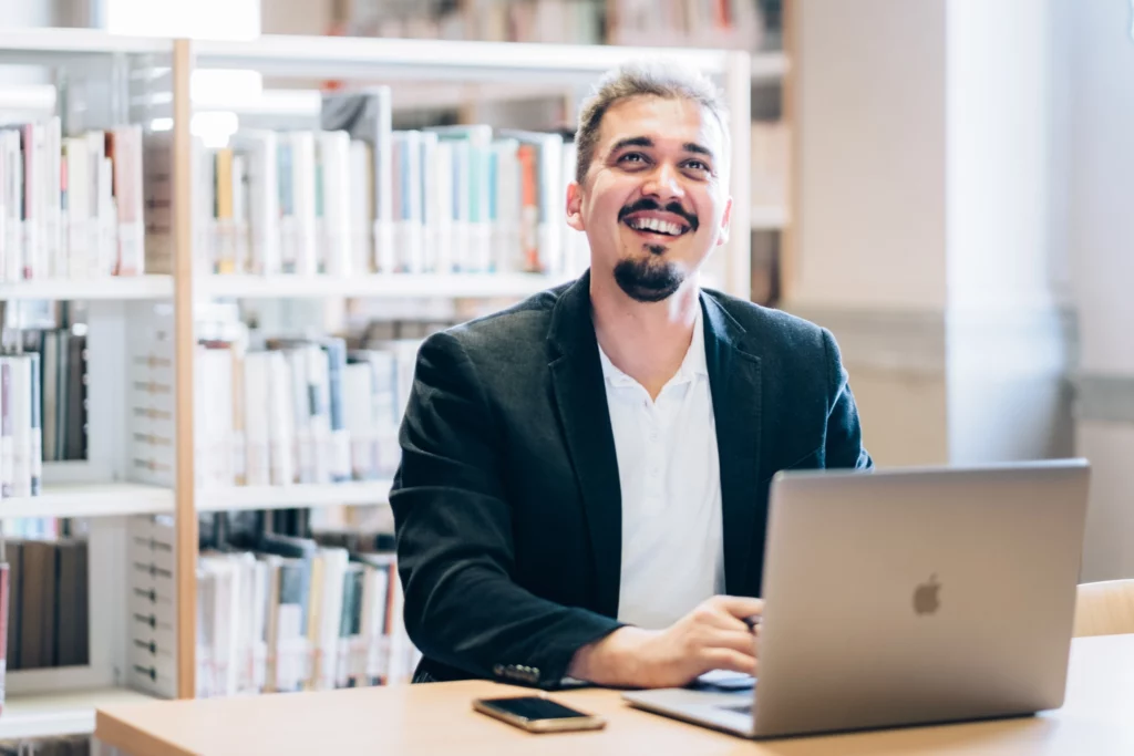 Kristian Selimi - Fondatore di Valutenze.it Kristian Selimi al lavoro con un laptop in una libreria, sorridente, in abito professionale.