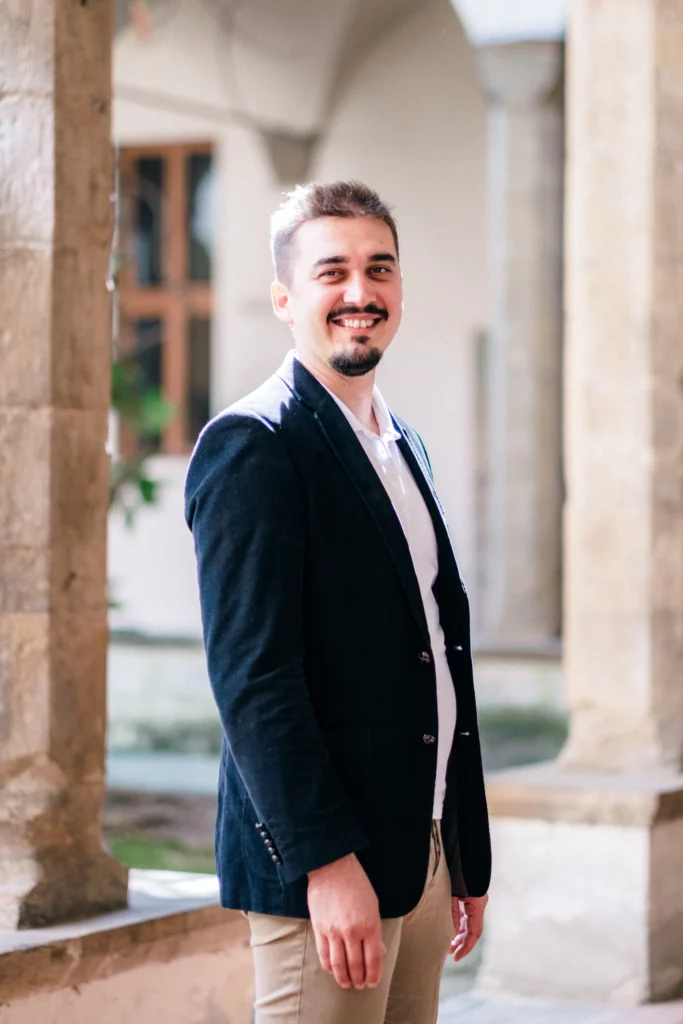 Kristian Selimi - Fondatore di Valutenze.it Kristian Selimi in piedi all'aperto, sorridente, in abbigliamento formale.